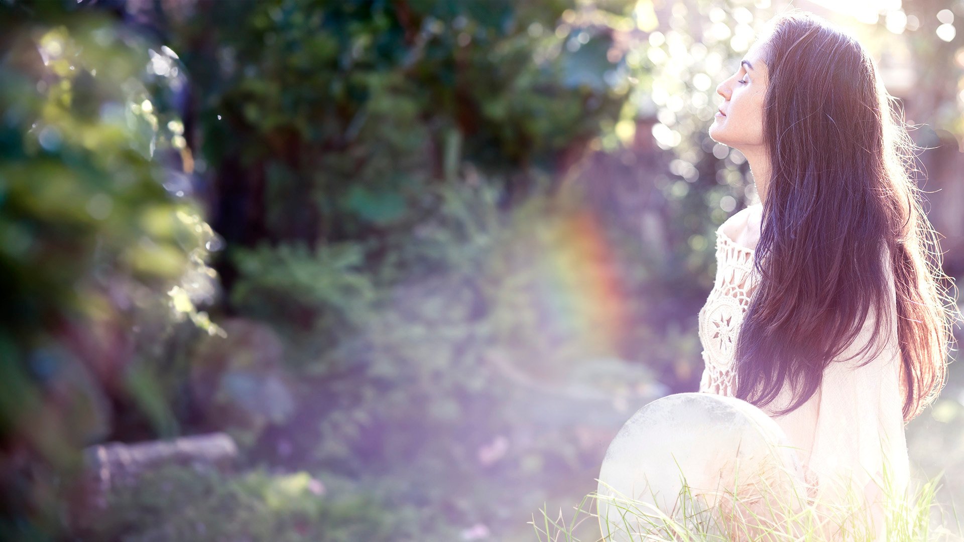 Sound Healing Hawaii, Reiki, Holistic Wellness - Sound Healer holding a frame drum sitting in the grass with a rainbow and sparkly trees in the background